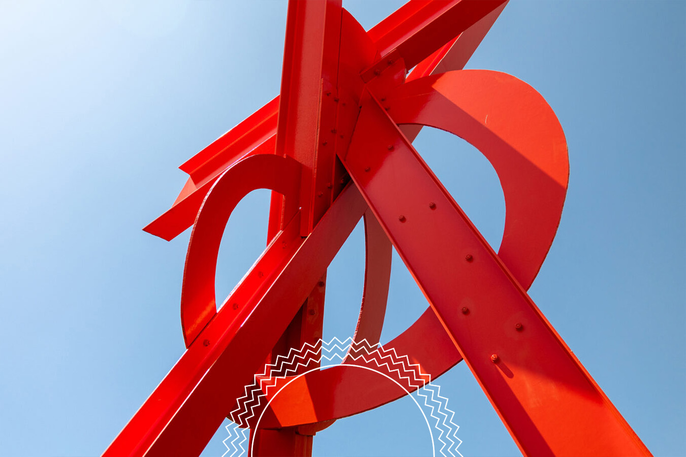 Red metal abstract sculpture with intersecting beams and curves, viewed from below against a clear blue sky.