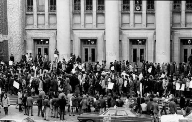 A large crowd of people with signs gathers in front of a building with columns, as “STRIKE” is written on the doors; a car is parked in the foreground.
