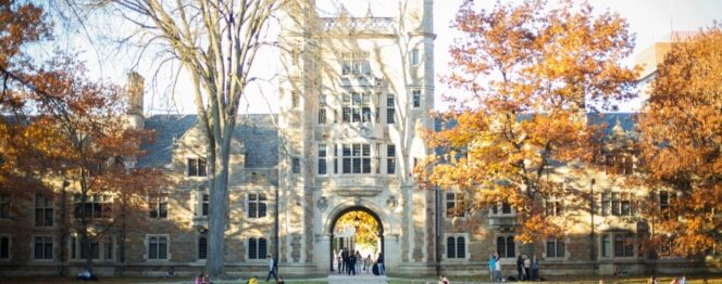 Stone university building with a large arched entrance, surrounded by autumn trees; people are walking and sitting on the lawn.