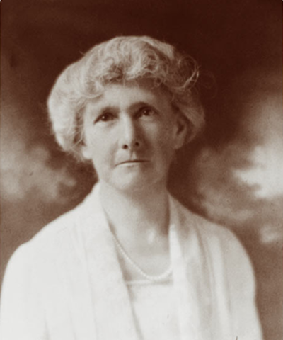 Sepia-toned portrait of an older woman with short, wavy hair, wearing a light blouse and a pearl necklace, looking slightly upward against a blurred background.