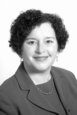 A woman with short curly hair, wearing a suit jacket, necklace, and earrings, smiles at the camera against a plain background.