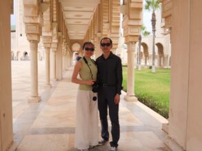 A couple posing together under a covered outdoor walkway with columns and arches, with grass and palm trees visible in the background.