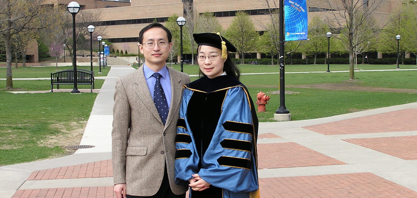 Two people stand on a brick walkway outside a university building; one wears academic regalia, and the other is dressed in a suit.