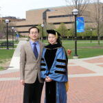 Two people stand on a brick walkway outside a university building; one wears academic regalia, and the other is dressed in a suit.