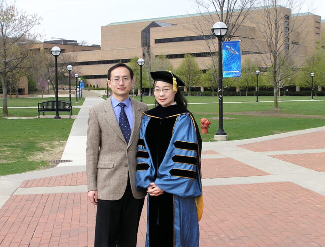 Two people stand on a brick walkway outside a university building; one wears academic regalia, and the other is dressed in a suit.