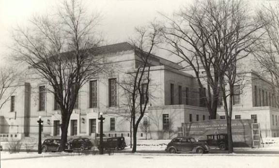 Black and white photo of a large stone building with tall windows, leafless trees in front, several vintage cars parked, and snow covering the ground.