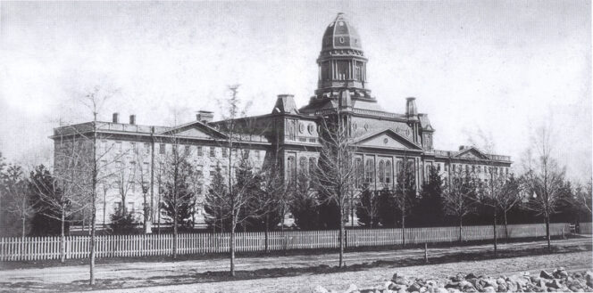 Black and white photo of a large domed building with trees, a white picket fence, and a rocky foreground.