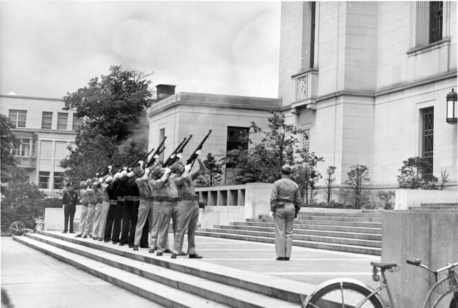 A firing squad of uniformed men performs a rifle salute on the steps outside a large building, with smoke visible from the rifles. A bicycle is parked in the foreground.