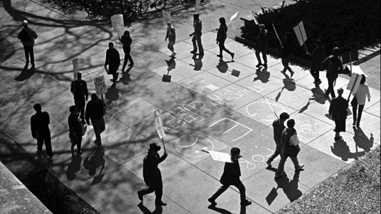 A group of people holding signs walk in a circular formation on a sidewalk with chalk writings visible on the ground.