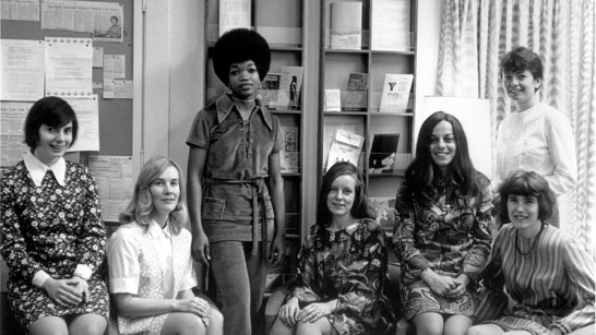 Seven women pose in an office setting, with bookshelves and a bulletin board in the background. Some are seated while others stand, all dressed in 1960s or 1970s-style clothing.