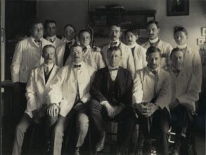 A group of twelve men in lab coats and formal attire pose for a formal portrait.