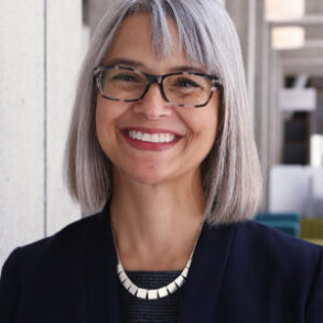 A smiling woman with shoulder-length gray hair, wearing glasses and a dark blazer.