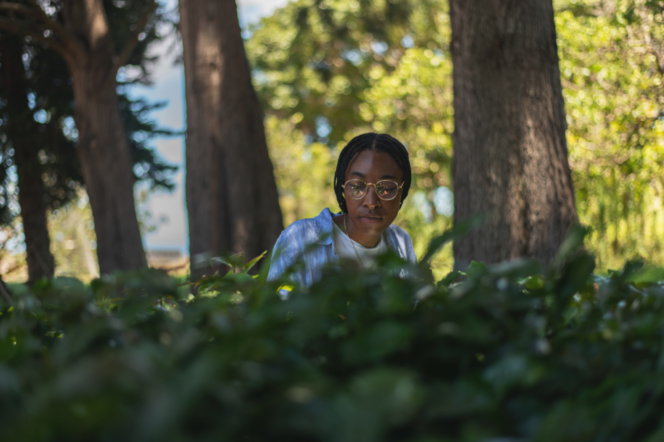 A person with braided hair and glasses looks down while sitting behind a hedge in a park with trees.