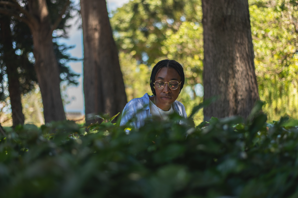 A person with braided hair and glasses looks down while sitting behind a hedge in a park with trees.