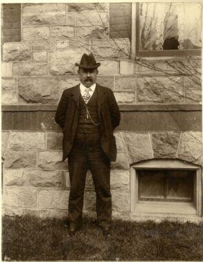 A man with a mustache, wearing a suit, vest, and fedora, standing before a stone building.