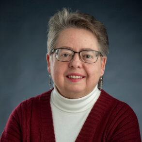A woman with short gray hair and glasses wearing a white turtleneck and red cardigan.