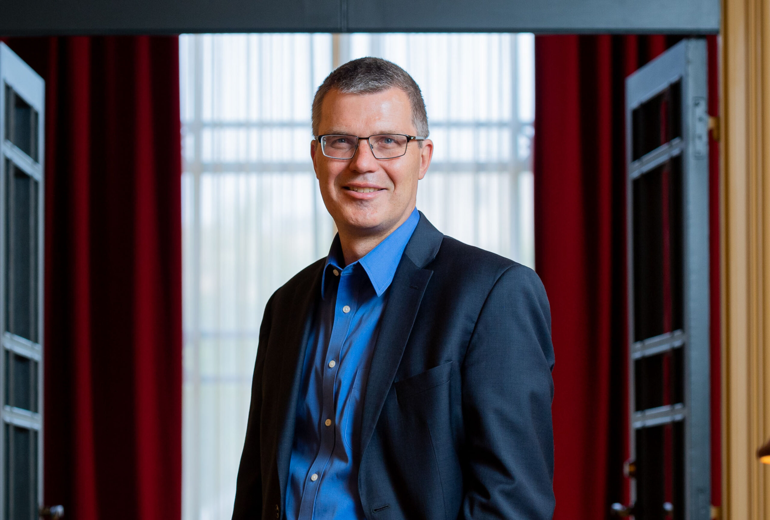 Middle-aged man wearing glasses, a blue button-down shirt, and a dark blazer standing in front of a window with red curtains.